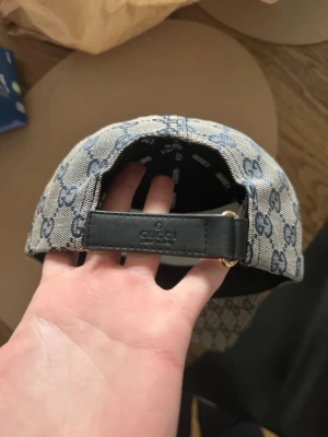  keps med GG-mönster - Snygg keps  i beige tyg med blått GG-monogram över hela kepsen. Justerbar rem bak i svart läder och klassisk -rand i rött och blåt på sidan. Perfekt för dig som vill ha en exklusiv och trendig accessoar.