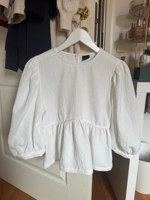 Vit blus med puffärm - Säljer en vit blus med puffiga långärmar och rund hals. Blusen har en strukturerad, lätt krinklad yta och en lös, flowy passform med avskärning i midjan. Stängs med knapp i nacken. Perfekt för en trendig och stilren look.