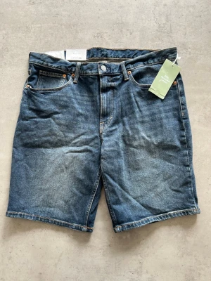 Blå denimshorts - W36 - Säljer ett par klassiska blå denimshorts med fem fickor och knappstängning. Shortsen har en rak passform och är tillverkade i bomull. Perfekta för sommarens alla äventyr, snygga att matcha med t-shirt eller skjorta.