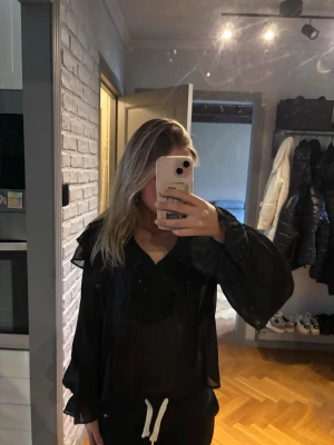 Svart transparent blus med volanger - Snygg svart blus i transparent material med volangdetaljer och v-ringning. Blusen har långa, luftiga ärmar och en avslappnad passform. Perfekt för dig som gillar en trendig och luftig stil.