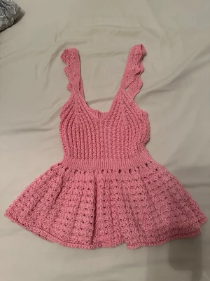 Rosa stickad linnetopp med volang - Supersöt rosa stickad linnetopp med volangdetaljer på axelbanden och v-ringad hals. Toppen har en figurnära passform upptill och en utställd, luftig nederdel. Perfekt för dig som vill ha en unik och feminin look.