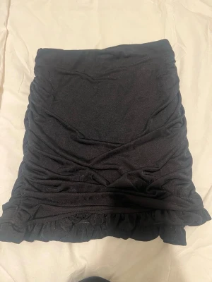 Svart kortkjol med volangkant - Snygg svart kortkjol med rynkade sidor och volangkant nedtill. Kjolen har en tight passform och är perfekt för att skapa en trendig look. Tillverkad i mjukt material som känns bekvämt mot huden.