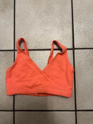 Lily of France orange sport-bh, storlek 34 - En mjuk och bekväm bh i korallfärgad bomull med omlott-design framtill och breda axelband. Bh:n har ingen bygel och ger ett naturligt stöd, perfekt för dig som vill ha en enkel och stilren look. Passar bra under toppar och tröjor.