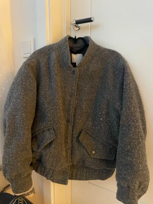 Grå bomberjacka från H&M - Snygg grå bomberjacka från H&M i ullmix med mjuk, lite luddig yta. Jackan har ribbade muddar, två fickor med lock och tryckknapp samt klassisk bomberkrage. Bra skick!