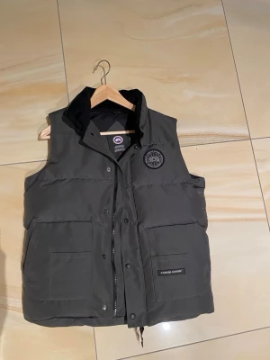 Mörkgrå dunväst från Canada Goose - Snygg mörkgrå dunväst från Canada Goose med hög krage och två stora fickor framtill. Västen har dragkedja och tryckknappar samt en broderad logga på bröstet. Perfekt för lager på lager och riktigt varm under kalla dagar.