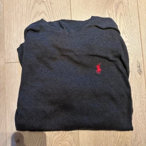 Mörkgrå stickad tröja från Ralph Lauren - Mörkgrå stickad tröja från Ralph Lauren med klassisk röd broderad logga på bröstet. Tröjan har v-ringad hals och är perfekt för dig som gillar stilren och tidlös design. Passar lika bra till jeans som till chinos.