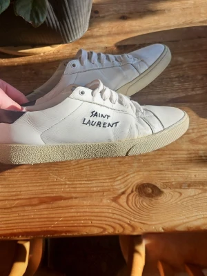 Vita Saint Laurent sneakers - Säljer ett par klassiska vita sneakers från Saint Laurent med handskriven logga på sidan. Skorna har låga skaft, vita snören och en beige sula med textur. Tillverkade i slätt läder för en clean och stilren look.