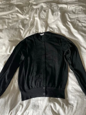 Svart klassisk kofta med knappar från Filippa k  - En stilren svart kofta med rund halsringning och knäppning framtill. Tillverkad i ett tunt, mjukt stickat material som passar perfekt till lager på lager. Långa ärmar och ribbade muddar vid ärmslut och nederkant ger en snygg passform.