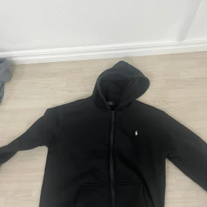 Tracksuit  från Polo Ralph Lauren - Säljer en svart hoodie från Polo Ralph Lauren med dragkedja framtill och klassisk huva. Tröjan har den ikoniska vita logon broderad på bröstet och fickor på sidorna. Perfekt för en avslappnad och stilren look.