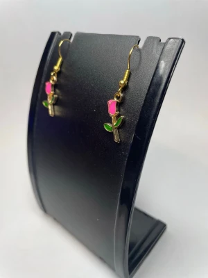 Hängande örhängen med rosor - Säljer ett par söta örhängen i form av små rosor. De har guldfärgade krokar och hängen med rosa blomma och gröna blad. Perfekta för dig som vill ha en romantisk och lekfull touch till din stil.