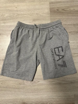Grå shorts från Emporio Armani EA7 - Snygga grå shorts från Emporio Armani EA7 med tryckt logga på vänster ben. Shortsen har elastisk midja med snörning och sidofickor med dragkedja. Perfekta för en avslappnad och sportig stil.