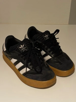 Adidas svartvita sneakers med gummisula - Snygga Adidas sneakers i svart läder med klassiska vita ränder på sidorna och vit häl. Skorna har svart snörning och en retro-inspirerad brun gummisula. Storlek US 5, priser är hej hugget i sten. Hör gärna av vid intresse, mvh Erik.