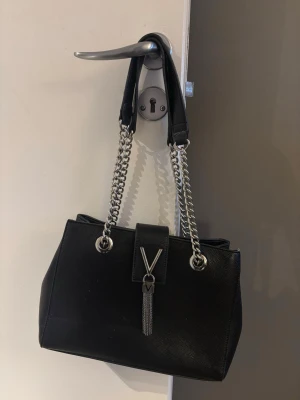 Svart handväska med kedjedetaljer - Snygg svart handväska från Valentino med silverfärgade kedjehandtag och dekorativt V-hänge med tofs framtill. Väskan har en stilren, rektangulär form och är tillverkad i skinnliknande material. Perfekt för dig som vill ha en trendig accessoar med lyxig känsla.