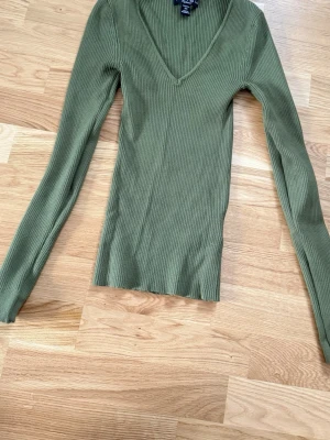 Olivgrön ribbstickad tröja Amisu - Snygg olivgrön ribbstickad tröja från Amisu med djup V-ringning och långa ärmar. Tröjan är figurnära och har en mjuk, stretchig känsla som passar perfekt till jeans eller kjol. Materialet är tunt och följsamt, vilket gör den enkel att styla.