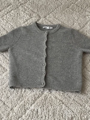 Grå kort kofta från Zara - Kort grå kofta från Zara med rund halsringning och pärlknappar framtill. Koftan är stickad i mjukt material och har ribbade muddar vid ärmslut och nederkant. Perfekt att slänga över en topp för en chill look.