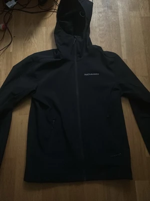 Svart vindjacka från Peak Performance - Snygg svart vindjacka från Peak Performance med huva och hel dragkedja framtill. Jackan har två sidofickor med dragkedja och diskret logga på bröstet. Använd en skidresa. Nypris 3000. Storlek M. Kan mötas upp i Stockholm eller skicka 