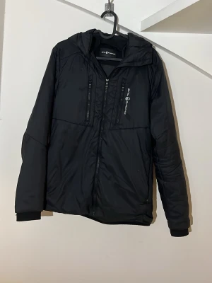 Svart vindjacka från Sail Racing - Svart vindjacka från Sail Racing med huva och två bröstfickor med dragkedja. Jackan har en sportig design och logotyp på bröstet. Materialet är syntet och jackan har ribbade muddar vid ärmslut. Perfekt för blåsiga dagar.