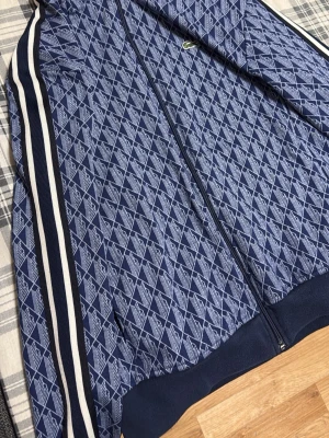 Blå  hoodie från Lacoste -   blå hoodie från Lacoste med vita ränder längs ärmarna.  Storlek passar för er som bär storlek M