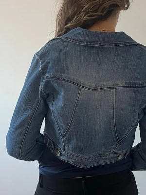Kort blå jeansjacka - Snygg kort jeansjacka i klassisk blå denim med markerade sömmar och silverfärgade knappar baktill. Jackan har en figurnära passform och tidlös design som passar perfekt till många olika outfits.