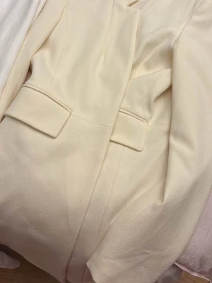 Krämvit klassisk kappa - Stilren krämvit kappa med raka linjer och två stora ficklock framtill. Kappan har en elegant, minimalistisk design och är tillverkad i ett mjukt tyg som ger en exklusiv känsla. Perfekt för dig som gillar tidlös och chic stil.