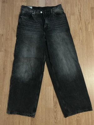 Baggy svarta jeans från H&M - Baggy jeans från H&M i tvättad svart färg med bred passform och raka ben. Jeansen har klassisk femficksdesign och är tillverkade i bomull. Perfekta för en avslappnad och trendig stil.