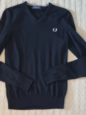 Mörkblå stickad tröja Fred Perry - Snygg mörkblå stickad tröja från Fred Perry med V-ringning och den klassiska vita lagerkransen broderad på bröstet. Tröjan har långa ärmar och ribbade muddar vid ärmslut och nederkant. Perfekt till jeans eller chinos. Fint skick använd fåtal ggr. 