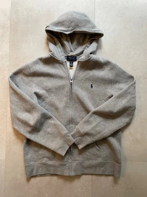 Grå hoodie från Polo Ralph Lauren - Säljer en grå hoodie från Polo Ralph Lauren i storlek S. Hoodien har dragkedja framtill, två fickor och en broderad logga på bröstet. Tillverkad i mjukt material med borstad insida för extra komfort. Perfekt för en avslappnad och stilren look.