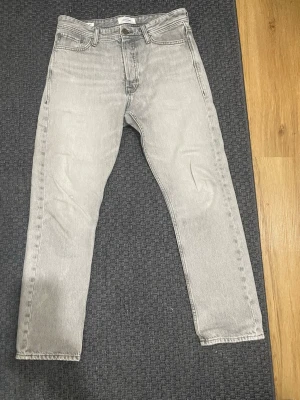 Grå relaxed jeans från Jack & Jones - Säljer ett par ljusgrå relaxed fit jeans från Jack & Jones, modell Chris. Klassisk femficksdesign och raka ben. Jeansen är tillverkade i denim och har en avslappnad passform som passar perfekt till en casual stil.