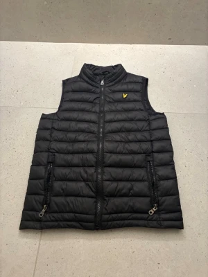 Svart dunväst från Lyle & Scott - Svart dunväst från Lyle & Scott med gul broderad logga på bröstet. Västen har ståkrage, två dragkedjefickor och quiltad design. Perfekt för lager-på-lager och enkel att matcha med olika outfits.