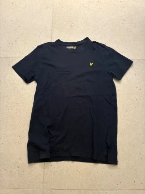 Svart t-shirt från Lyle & Scott - Klassisk svart t-shirt från Lyle & Scott med rund hals och korta ärmar. På bröstet sitter den ikoniska gula örnloggan broderad. Tillverkad i mjuk bomull som känns skön mot huden. Perfekt basplagg för en clean och stilren look.