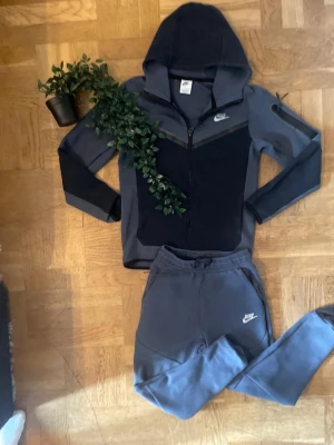 Nike tech med både byxor/hoddie - Nike loungewear set i blågrå färg med matchande huvtröja och byxor. Tröjan har dragkedja, huva och Nike-logga på bröstet. Byxorna har resår i midjan och Nike-logga på låret. Perfekt för chill dagar hemma.