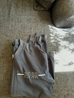 Mörkgrå loungewear-set från Hugo Boss - Snyggt mörkgrått loungewear-set från Hugo Boss med dragkedja framtill och två fickor med dragkedja. Setet har diskret logga på bröstet och är tillverkat i mjukt material, perfekt för chill dagar hemma.