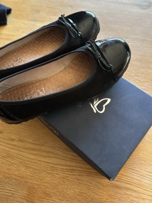 Svarta ballerinaskor från Caprice - Nypris 1000kr. Stilrena svarta ballerinaskor från Caprice med lackad tå och liten rosett framtill. Skorna har en mjuk innersula med prickig struktur för extra komfort. Klassisk rundad form och diskret logga på innersulan.