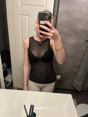 Svart spetsig ärmlös linnetopp - Säljer en svart, genombruten spetslinnetopp med snyggt mönster. Toppen är ärmlös och har en tight passform som framhäver figuren. Perfekt att styla med jeans eller kjol för en trendig look.