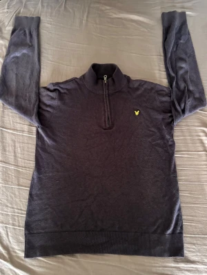 Mörkgrå halvzip-tröja från Lyle & Scott - Snygg mörkgrå stickad tröja från Lyle & Scott med halvzip och ståkrage. Tröjan har ett gult broderat logomärke på bröstet och ribbade muddar vid ärmslut och nederkant. Perfekt för en stilren och avslappnad look. Storleken: 152/158(12-13 years)