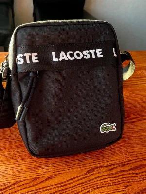 Svart axelremsväska från Lacoste - Snygg svart axelremsväska från Lacoste med tydlig logga framtill och ett band med Lacoste-tryck. Väskan har en fyrkantig form, dragkedja och justerbar rem. Perfekt för att bära det viktigaste och ger en sportig vibe.