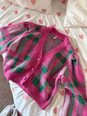 Rutig rosa och grön kofta - Säljer en färgglad kofta med rutigt mönster i rosa, grönt och ljusrosa. Koftan har v-ringning, ribbade muddar och tre klädda knappar framtill. 