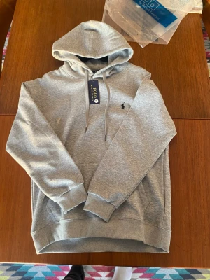 Grå hoodie från Polo Ralph Lauren - Snygg grå hoodie från Polo Ralph Lauren med klassisk huva och dragsnören. Tröjan har ribbade muddar vid ärmslut och nederkant samt den ikoniska lilla loggan broderad på bröstet. Tillverkad i mjukt bomullsmaterial för maximal komfort.
