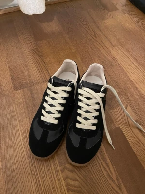 Maison Margiela skor - Maison Margiela sneakers i svart mocka och läder med vita kontrasterande snören och beige innersula. Klassisk låg modell med gummisula och stilren design. Kommer med originalkartong och dustbag.