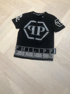 Svart Philipp Plein t-shirt med tryck - Svart t-shirt från Philipp Plein med stort PP-logotryck framtill, dekorativa detaljer på ärmarna och mönstrad kant längst ner med texten PHILIPP PLEIN. T-shirten har korta ärmar och rund hals. Materialet är mjuk bomull.