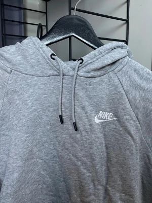 Grå hoodie från Nike med logga - Snygg grå hoodie från Nike med klassisk vit logga på bröstet. Hoodien har huva med dragsko och en stor magficka framtill. Tillverkad i mjukt sweatshirtmaterial som är perfekt för chill dagar. Enkel och stilren design som funkar till det mesta.