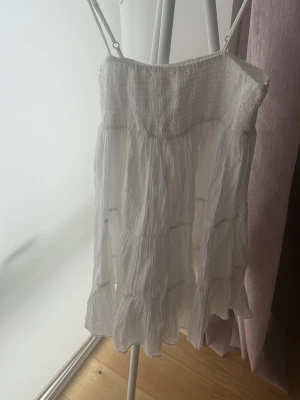 Vit somrig axelbandslös klänning - Luftig vit kortklänning med smock upptill och tunna axelband. Klänningen har volangdetaljer och är tillverkad i ett mjukt, skrynklat bomullstyg som ger en avslappnad känsla. Perfekt för varma dagar och sommarens alla tillfällen.skriv innan ni köper 