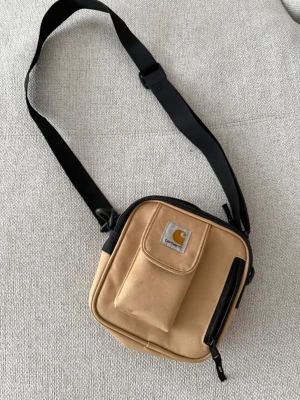 Beige axelväska från Carhartt - Snygg beige axelväska från Carhartt med svart justerbar rem. Väskan har en fyrkantig form, praktisk framficka med lock och dragkedja samt ett rymligt huvudfack. Tillverkad i slitstark canvas som ger en cool streetwear-vibe.