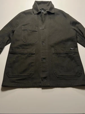 Grön overshirt från NN07 - Stilren olivgrön overshirt från NN07 i kraftig bomull. Klassisk krage, tre stora utanpåfickor och knäppning framtill. Perfekt lager-på-lager-plagg med avslappnad passform och diskret logolapp på ena fickan. Enkel och tidlös design som funkar till mycket.PASSAR SOM S. Pris kan diskuteras