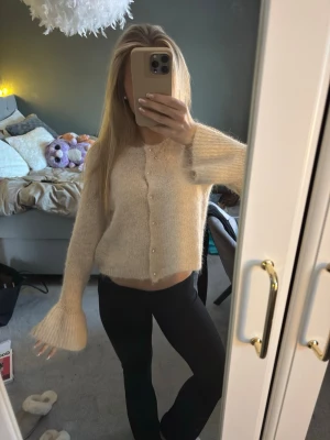 Beige kofta med pärlknappar - Supersöt beige kofta med fluffig känsla och pärlknappar framtill. Modellen är croppad med vida ärmar och mjukt material. Perfekt att styla med jeans eller kjol. Funkar till allt! Nästan oanvänd