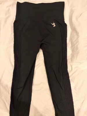 Svarta träningsbyxor från V3 Apparel - Säljer ett par svarta träningsbyxor från V3 Apparel med hög midja och ribbade detaljer längs sidorna. De har scrunch-effekt bak och är tillverkade i ett stretchigt material som sitter tight. Perfekta för träning eller chill.