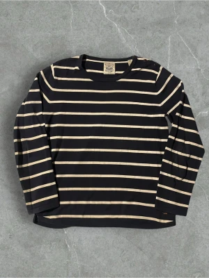 Randig marinblå tröja från Scoth & Soda - Säljer en klassisk långärmad tröja från Scotch & Soda i marinblått med vita horisontella ränder. Tröjan har rund halsringning och knappar på axeln för en snygg detalj. Tillverkad i mjuk bomull, perfekt för en avslappnad och stilren look.