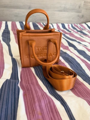 Ny brun mini handväska - Helt ny mini handväska i fin kamelbrun färg ✨ Oanvänd och i perfekt skick. Stilren modell med korta handtag samt justerbar & avtagbar axelrem. Perfekt storlek för mobil, plånbok och andra småsaker. Material: Faux leather Skick: Ny