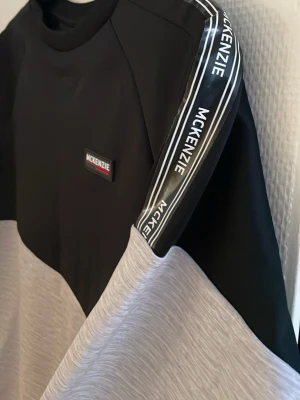 Svart och grå sweatshirt från McKenzie - Snygg sweatshirt från McKenzie i svart och ljusgrått med logga på bröstet och längs ärmarna. Tröjan har rund halsringning och raka muddar. Perfekt för en avslappnad streetwear-look. 13-15 år XL