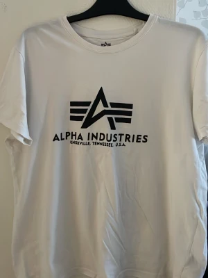 Vit Alpha Industries t-shirt - Snygg vit t-shirt från Alpha Industries med stor svart logga och text på bröstet. Klassisk rund halsringning och korta ärmar. Tillverkad i mjuk bomull som känns skön mot huden. Perfekt för dig som gillar streetwear och enkel stil.